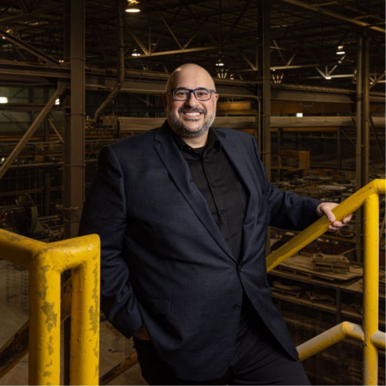salesperson Ernesto Polsinelli at the top of a staircase on a mezzanine inside a busy manufacturing plant. Machinery and workers in the background.