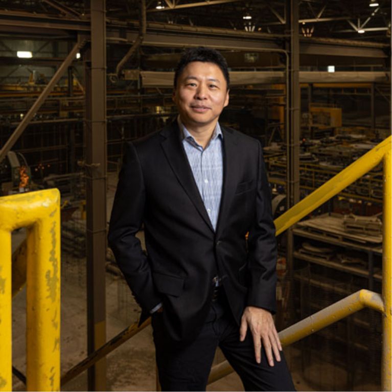 salesperson Barron Wang at the top of a staircase on a mezzanine inside a busy manufacturing plant. Machinery and workers in the background.